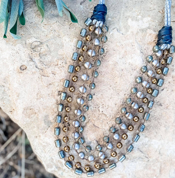 A multi-strand beaded necklace with metallic gold, silver, and pearl-like beads arranged on a rock surface, with a few green leaves visible in the top left corner.