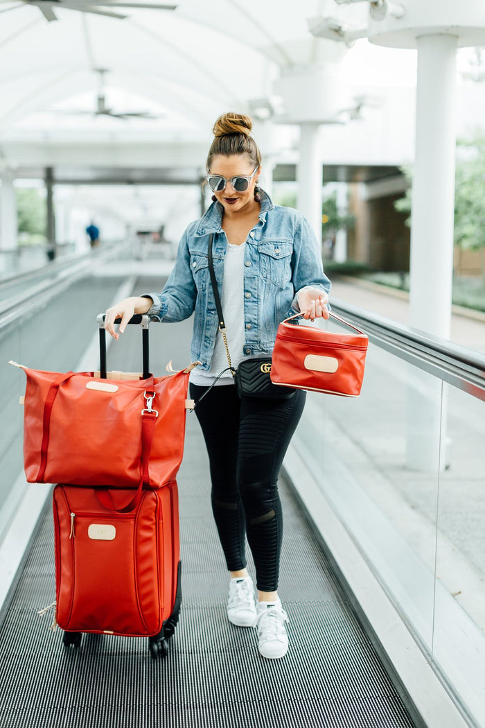 Woman in sunglasses and a denim jacket walks with a large red suitcase and matching red bag on an airport moving walkway. She wears black pants, white sneakers, and carries a small black purse.
