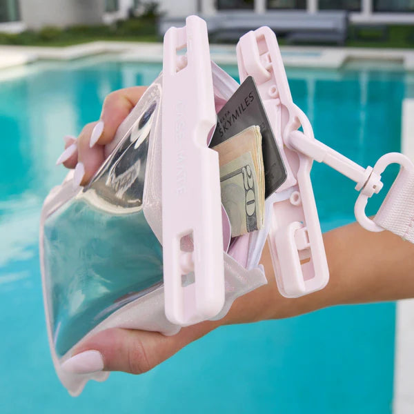 A hand holds the Ballet Pink Twinkle Wavy Waterproof Floating Phone Pouch with a phone, cash, and cards by a pool, water, and a modern building in the background.