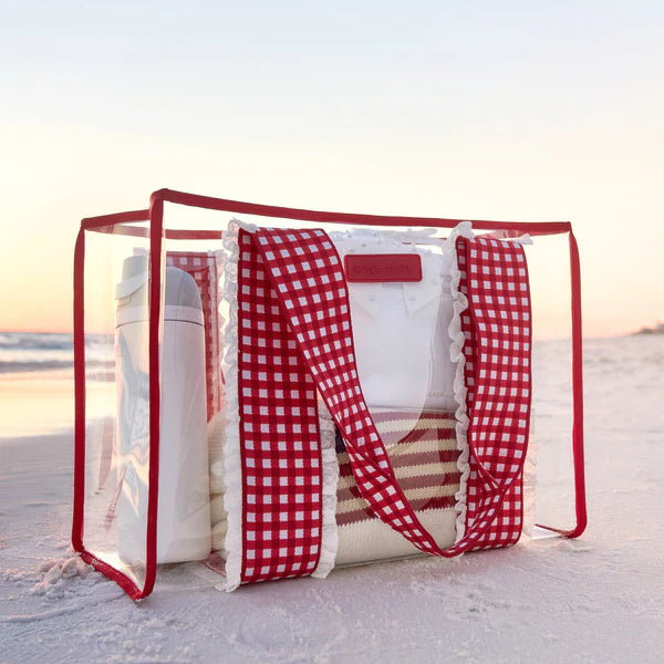 The Cherry Gingham Jelly Tote Bag W/ Phone Pouch rests on the sandy beach at sunset, holding a white water bottle and folded towels—an ideal summer travel companion, with the softly lit ocean and sky in the background.