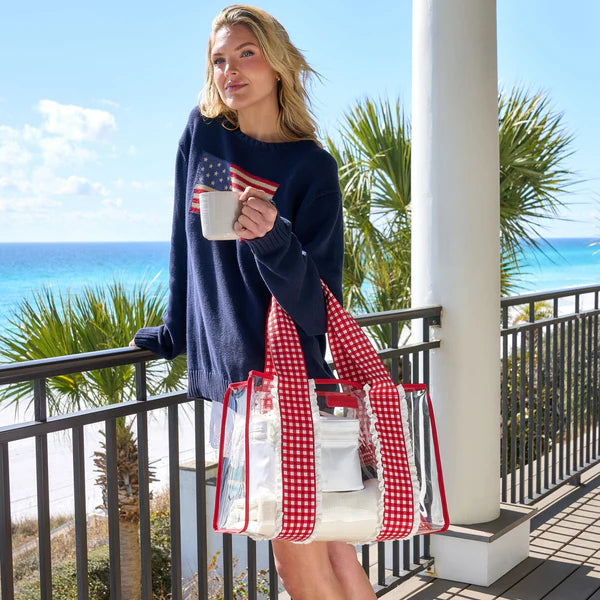 A woman in a navy sweater with an American flag design stands on a beachside balcony, holding her mug and Cherry Gingham Jelly Tote Bag W/ Phone Pouch—ideal for summer travel. Palm trees and the ocean are visible in the background.