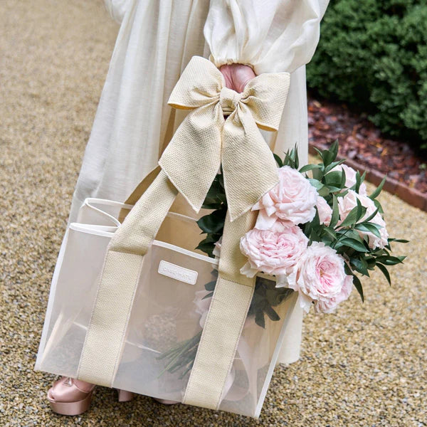 A person in a cream dress holds the Raffia Bow Jelly Tote Bag W/ Phone Pouch, featuring wide beige handles and a large bow, filled with pink peonies and greenery. They stand on a gravel path surrounded by lush foliage.