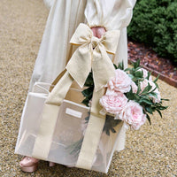 A person in a cream dress holds the Raffia Bow Jelly Tote Bag W/ Phone Pouch, featuring wide beige handles and a large bow, filled with pink peonies and greenery. They stand on a gravel path surrounded by lush foliage.