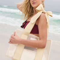 A woman on the beach holds the Raffia Bow Jelly Tote Bag W/ Phone Pouch, featuring wide beige straps and a large bow, against ocean waves and a cloudy sky—a chic accessory for stylish resort travel.