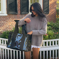 A woman with long dark hair, sunglasses, a taupe sweater, and a white skirt stands outdoors by a white fence, smiling as she holds the Midnight Black Bow Jelly Tote Bag W/ Phone Pouch. A brick building with shuttered windows is in the background.