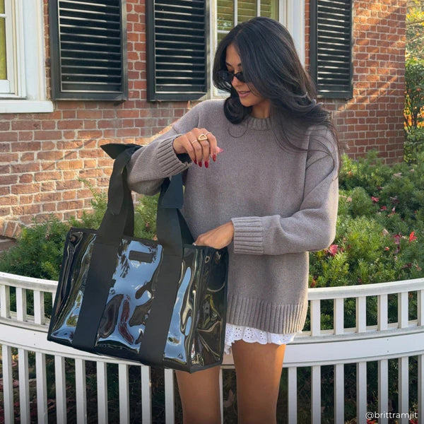 A woman with long dark hair, sunglasses, a taupe sweater, and a white skirt stands outdoors by a white fence, smiling as she holds the Midnight Black Bow Jelly Tote Bag W/ Phone Pouch. A brick building with shuttered windows is in the background.
